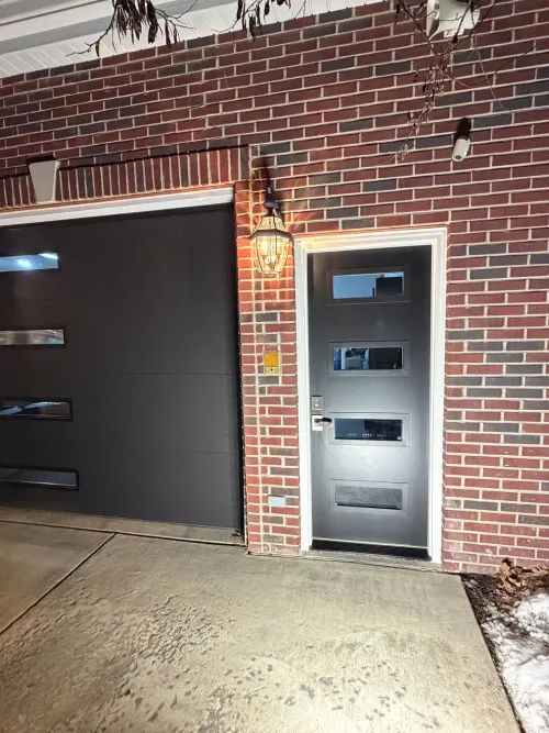 Black contemporary entry door alongside a matching black garage door on brick