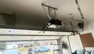 Ceiling-mounted garage door opener with wires and rails visible, technician outside inspecting tools near an open garage.