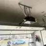 Ceiling-mounted garage door opener with wires and rails visible, technician outside inspecting tools near an open garage.